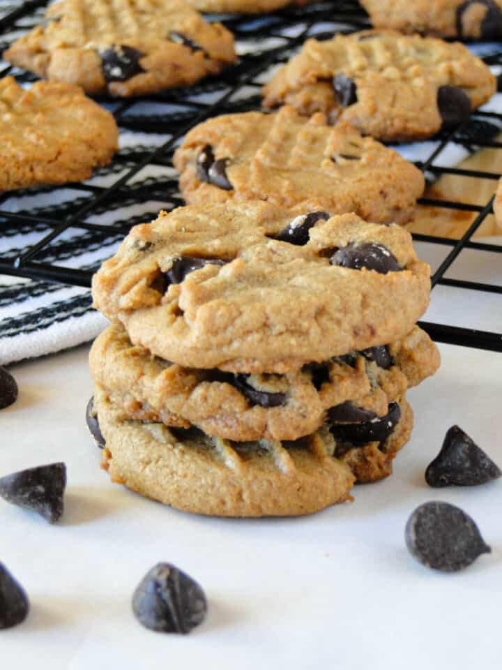 Three flourless peanut butter chocolate chip cookies stacked in front of cookies on wire rack.