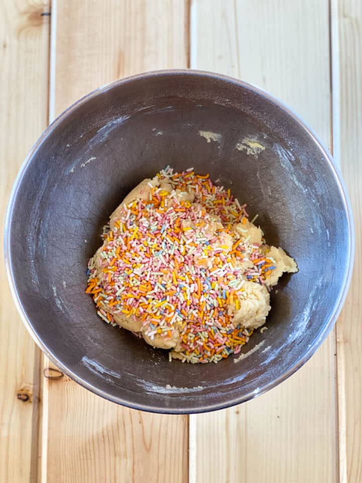 Rainbow sprinkles added to sugar cookie dough in large mixing bowl.