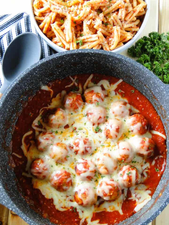 Chicken meatballs in sauce pan with sauce and melted cheese next to large white bowl of pasta.