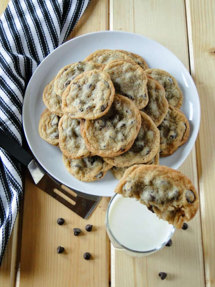 Classic chocolate chip cookies on white round plate behind a glass of milk with on cookie broken in half sitting on top of glass.