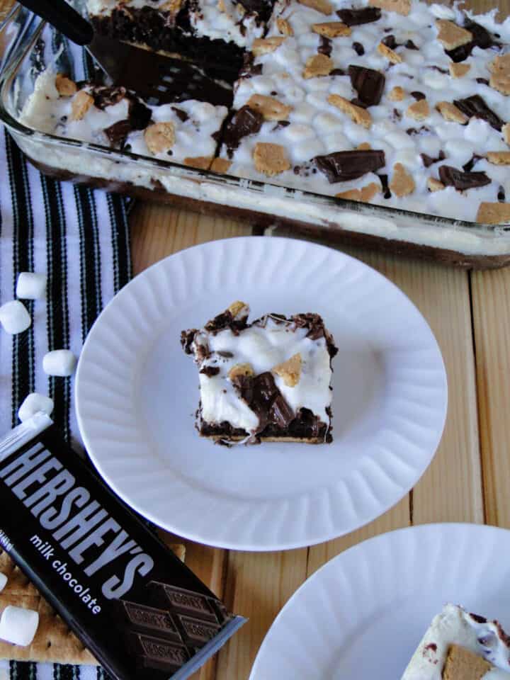Top view of smores brownie slice on white round plate in front of baking dish of brownies.