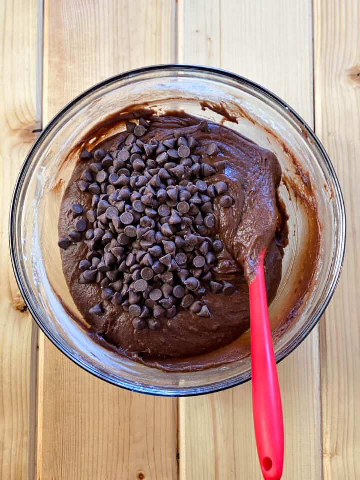 Brownie batter with chocolate chips on top in glass mixing bowl with red spatula.