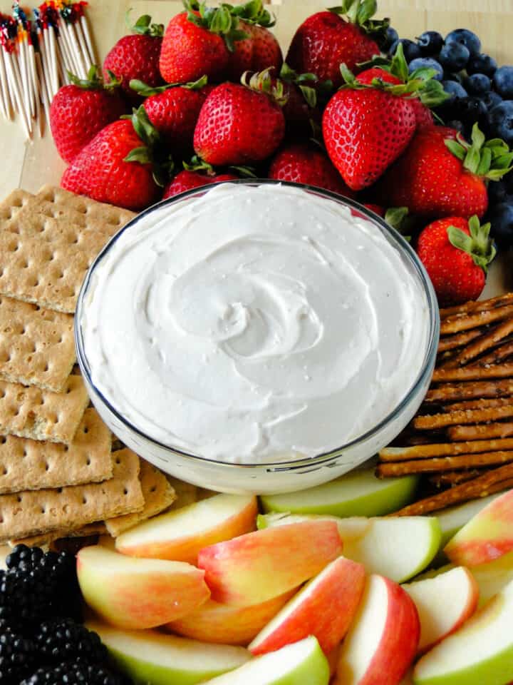 Yogurt fruit dip in bowl in center of board with fruit, graham crackers and pretzels around it.