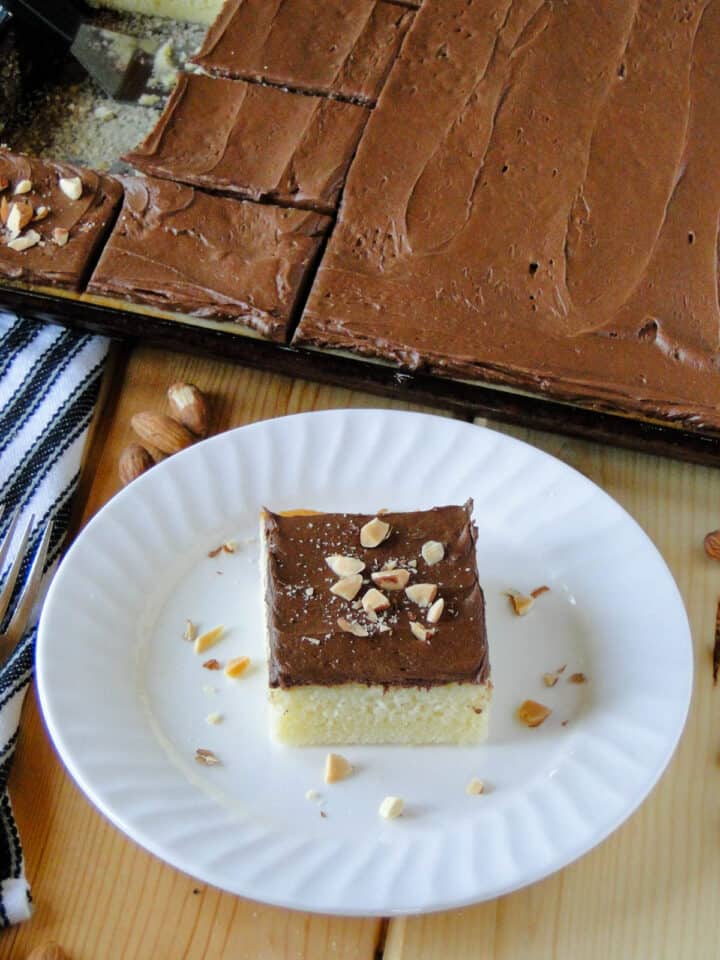 Slice of almond cake with chocolate frosting on white round plate in front of entire sheet cake.