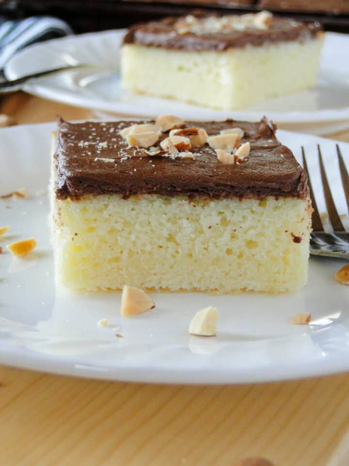 Close up side view of almond cake with chocolate frosting slice garnished with chopped almonds on white round plate with fork.