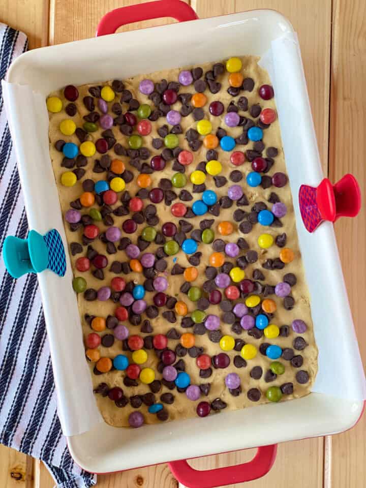 Chocolate chips and candies sprinkled on top of cookie dough in baking dish.