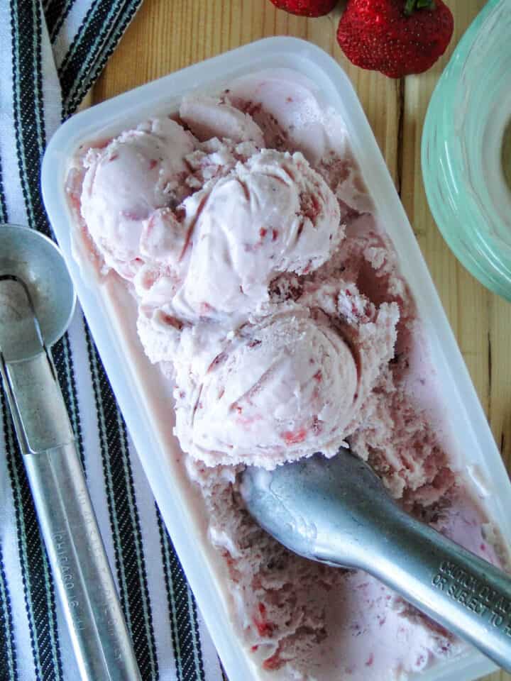 Container of homemade strawberry ice cream with scoops on top and ice cream scooper.