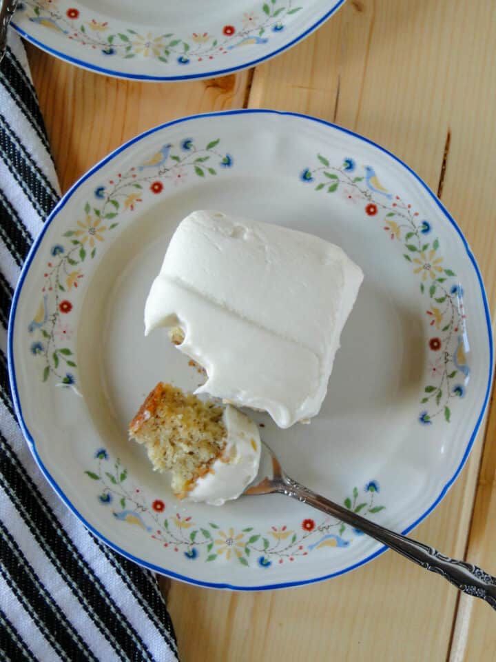 Top view of slice of banana cake with cream cheese frosting with bite on fork on white round plate.