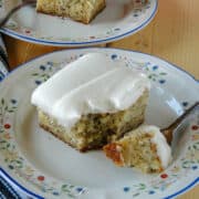 Close up side view of banana cake with cream cheese frosting slice with bite on fork on white round plate in front of another slice of cake on plate with fork.