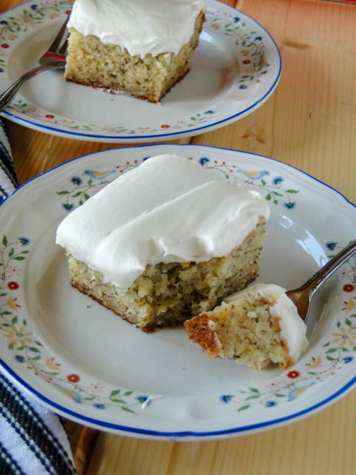 Close up side view of banana cake with cream cheese frosting slice with bite on fork on white round plate in front of another slice of cake on plate with fork.