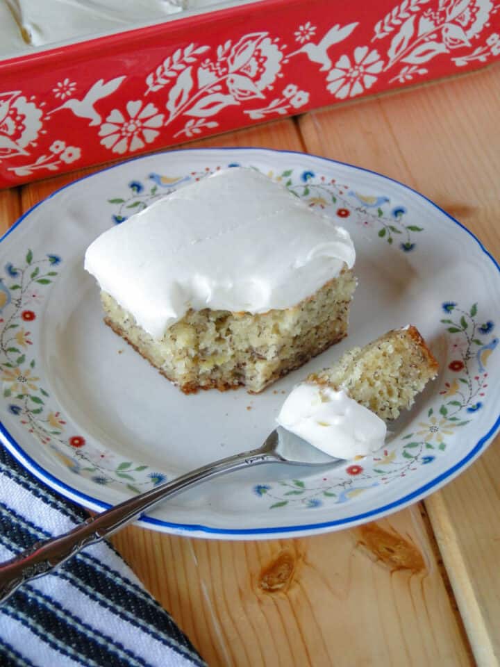 Slice of banana cake with cream cheese frosting with bite on fork on white round plate in front of pan of cake.