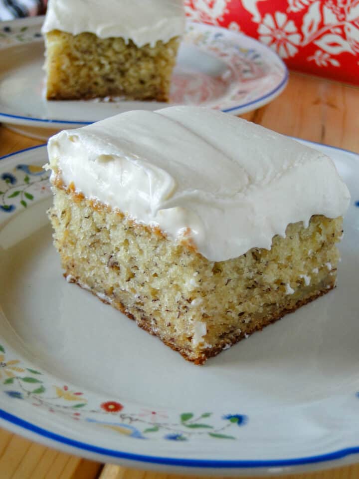 Close up side view of slice of banana cake with cream cheese frosting on white round plate in front of another slice of cake on plate.