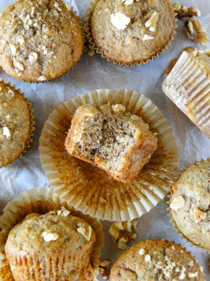 Close up top view of banana nut muffins with one muffin unwrapped on its side and a bite taken out of it.