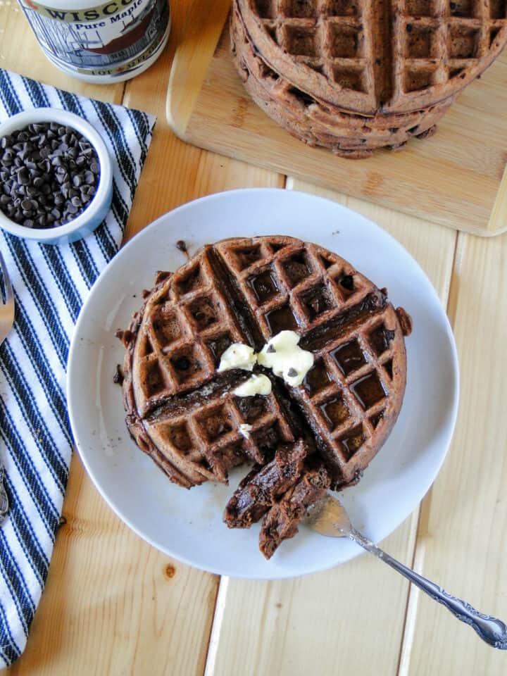 Chocolate waffles on white round plate with bite on fork in front of stack of waffles and small bowl of extra chocolate chips.