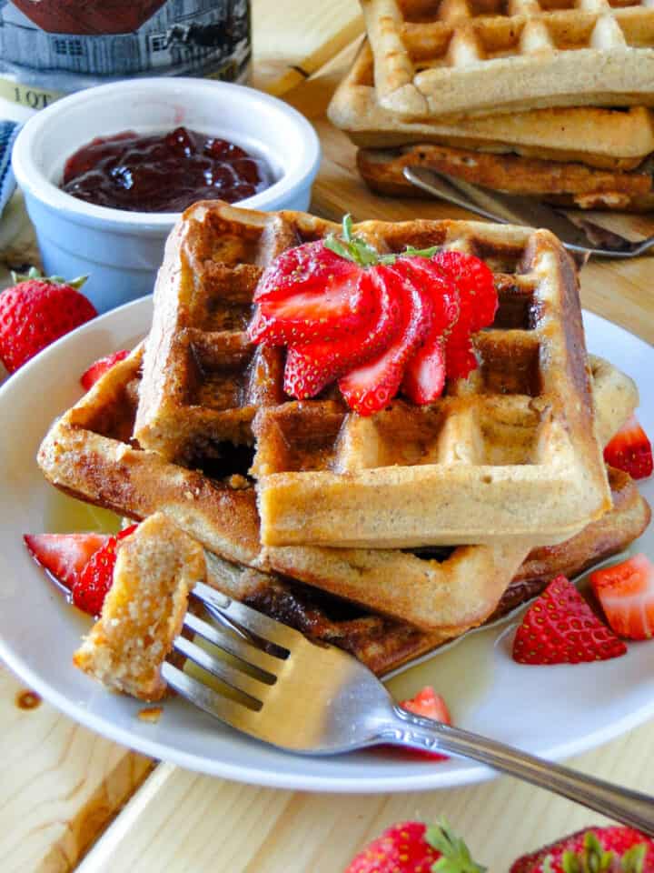 Peanut butter and jelly waffles stacked on white round plate with a strawberry fan on top and diced strawberries on the side with a bite of waffle on a fork.