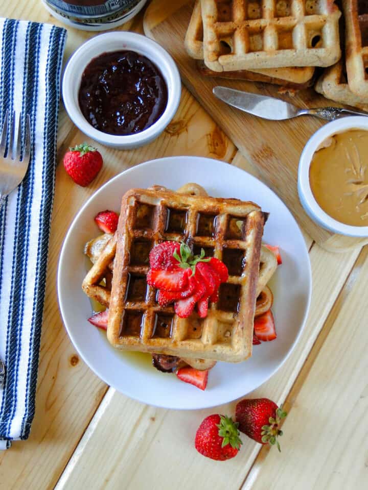 Peanut butter and jelly waffles stacked on a plate garnished with fresh strawberries and small bowls of peanut butter and jelly on the side.