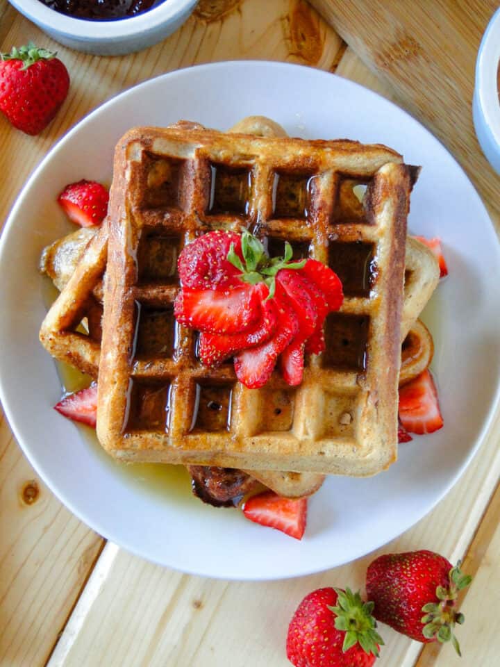 Top view of 3 peanut butter and jelly waffles stacked on white round plate and garnished with strawberry fan and diced strawberries and a drizzle of maple syrup.