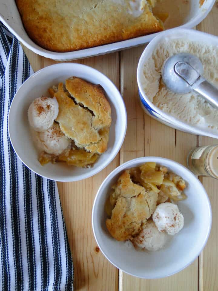 Two bowls of easy apple cobbler with ice cream in front of baking dish and tub of ice cream on the side with scooper.