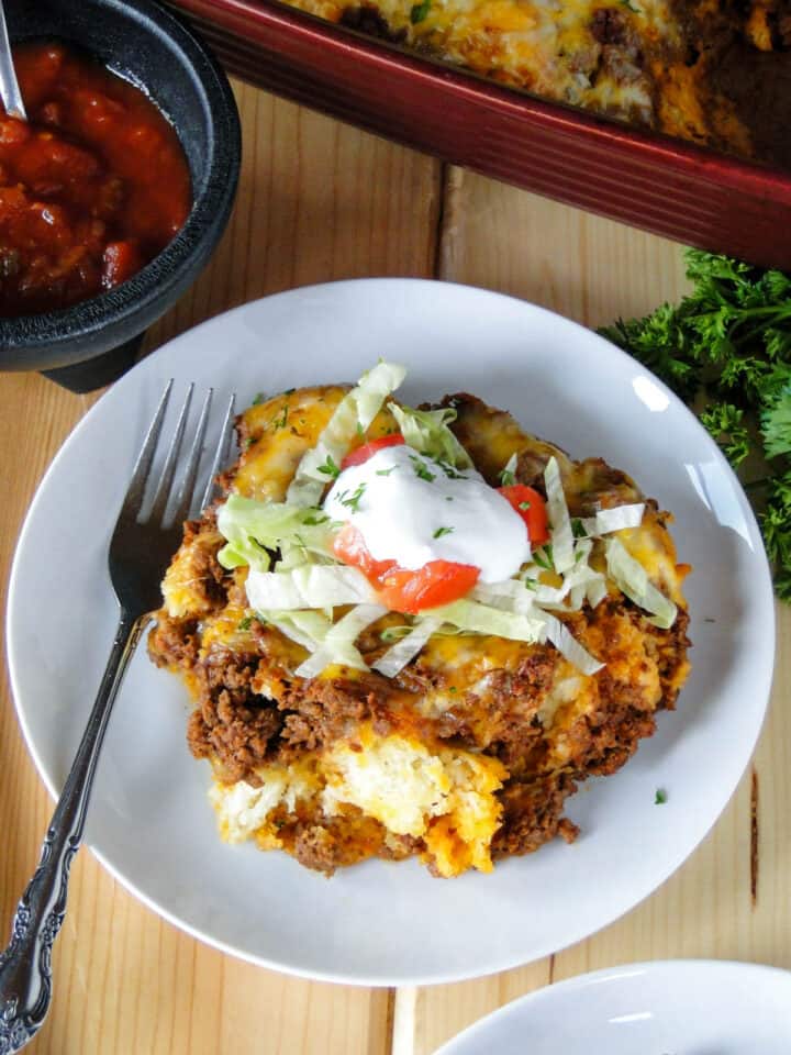 Taco biscuit bake on white round plate with fork in front of casserole dish.