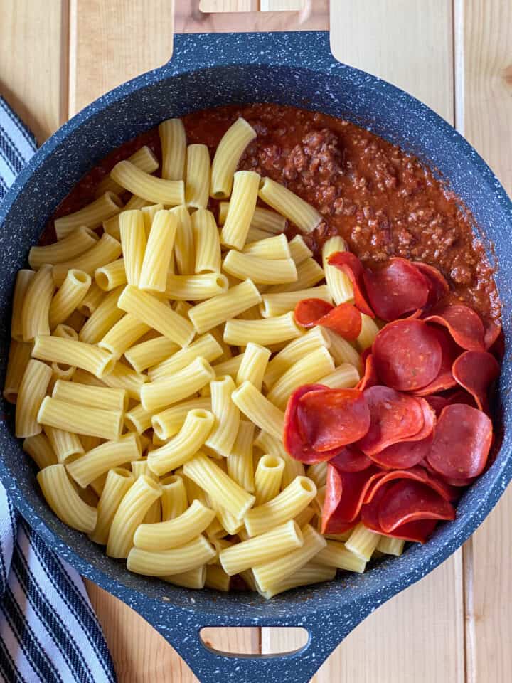 Cooked pasta and sliced pepperoni added to meat sauce in round skillet.