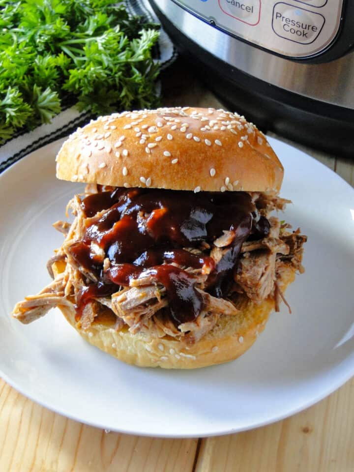 Pulled pork on bun with bbq sauce on white plate in front of instant pot.