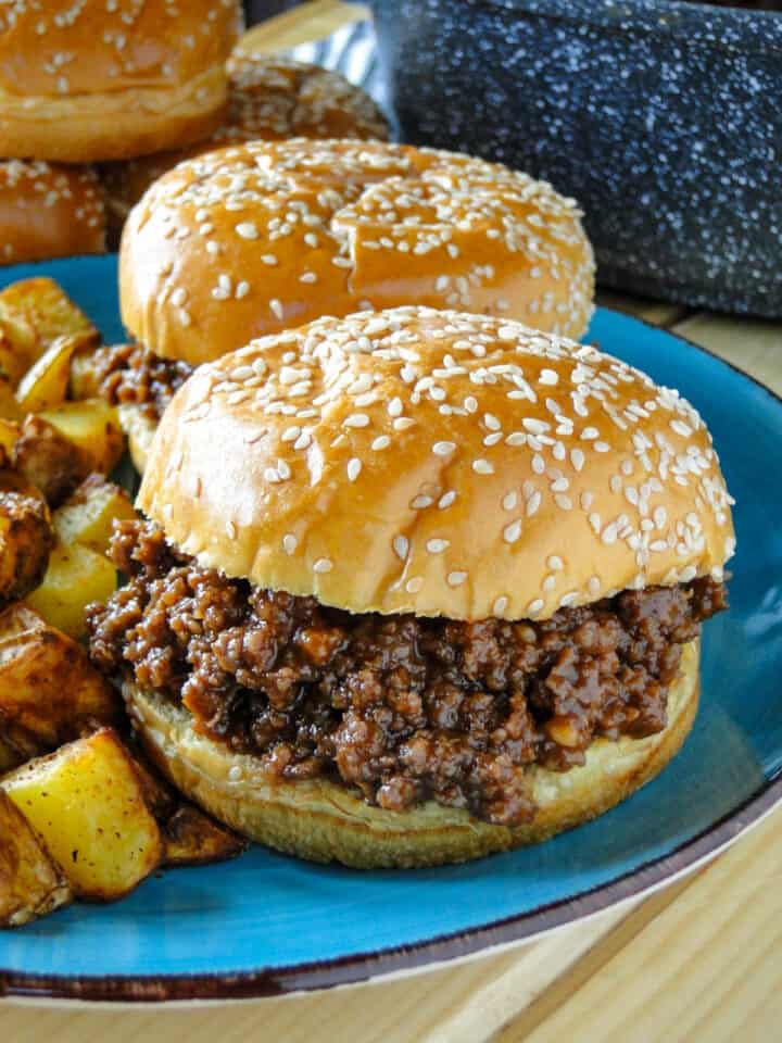 Side view of 2 easy homemade sloppy joes on blue plate in front of saucepan of sloppy joe mix and buns.