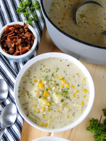 Creamy corn chowder in white round bowl in front of pot of soup and small bowl of bacon pieces on the side.
