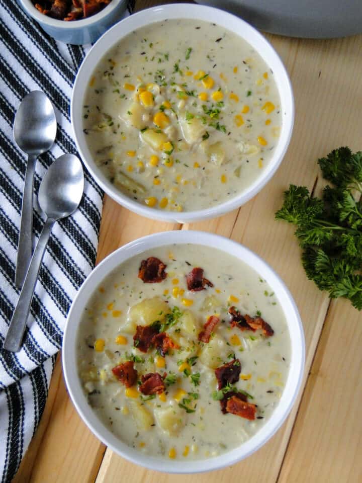 Two bowls of creamy corn chowder in white round bowls, one topped with bacon pieces, and two spoons on the side.