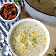 Creamy corn chowder in white round bowl in front of pot of soup and small bowl of bacon pieces on the side.