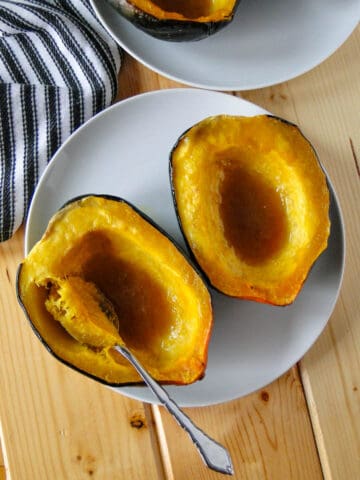 Maple roasted acorn squash halves on white round plate with spoon scooping out some squash.