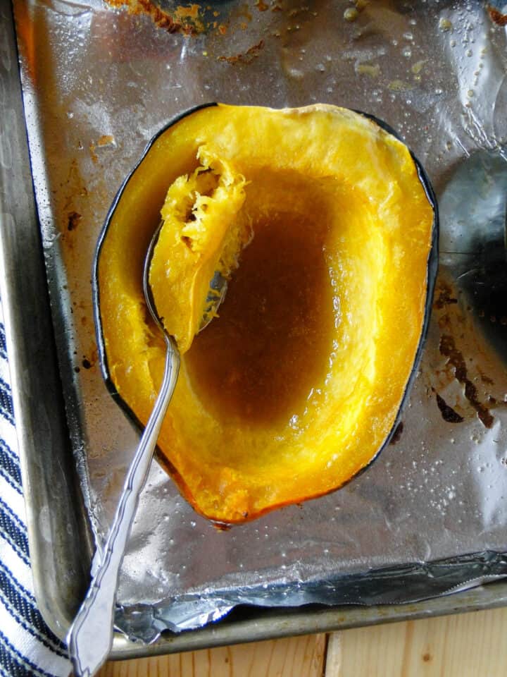 Close up view of maple roasted acorn squash half with spoon scooping out flesh on sheet pan.