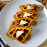 Top view of pumpkin waffle cut into quarters on white round plate with triangle pats of butter and maple syrup.