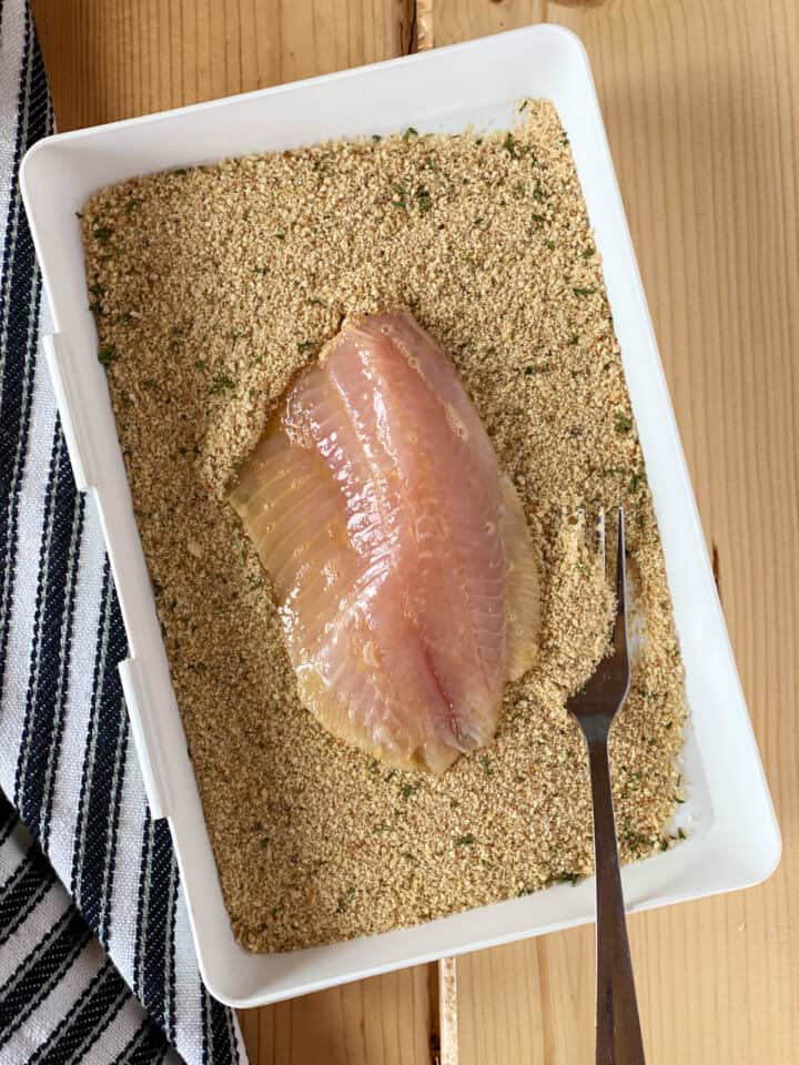 Egg coated tilapia filet siting in shallow dish of breading mixture with a fork.