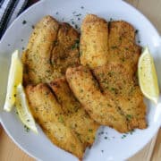 Air fryer breaded tilapia filets on white round plate with lemon wedges.