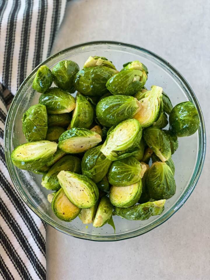Halved brussels sprouts in large glass mixing bowl coated with oil, salt and black pepper.
