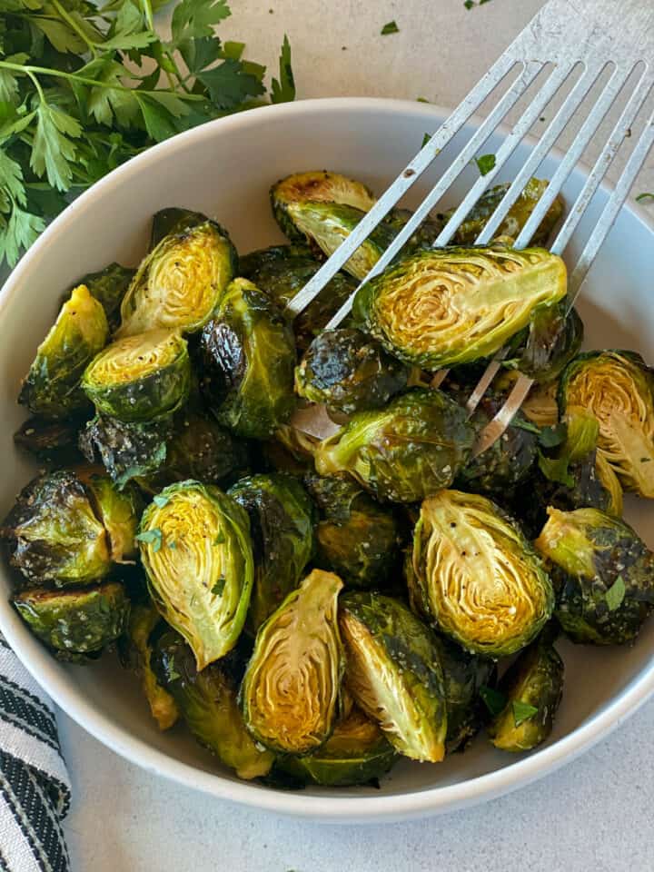 Roasted brussels sprouts in white round bowl with a few falling off a metal spatula in right side of bowl.