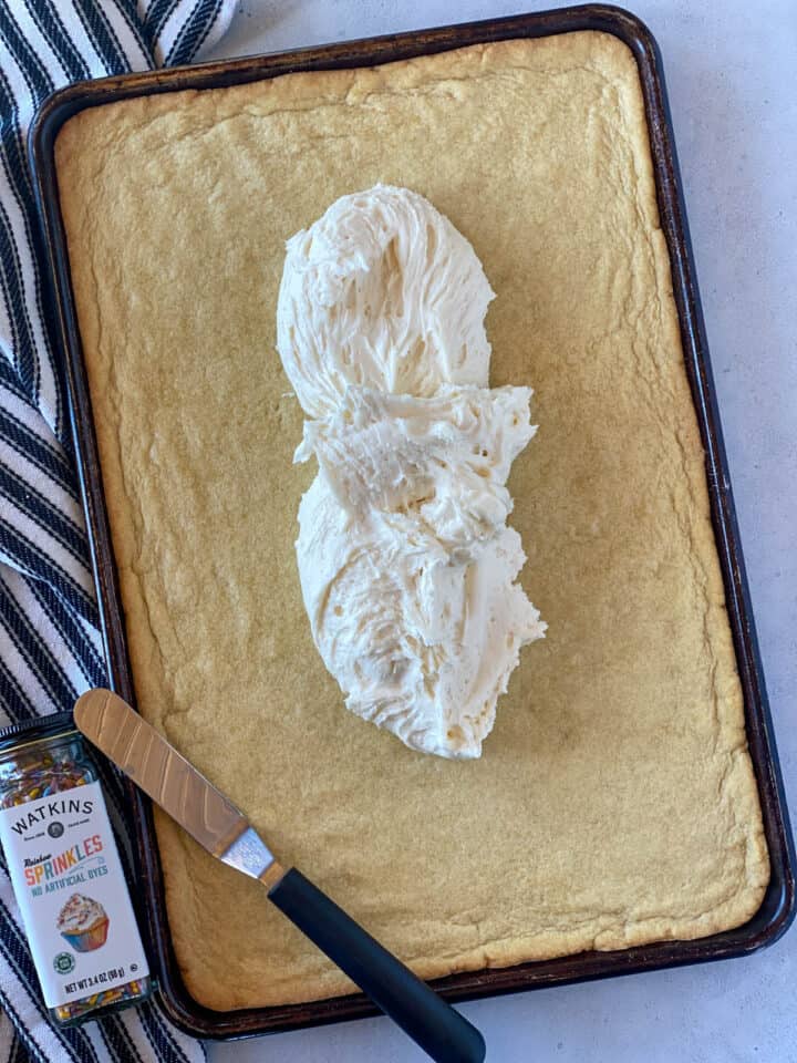 Buttercream frosting added to center of cookie bar in sheet pan with offset spatula and jar of sprinkles on the left side.