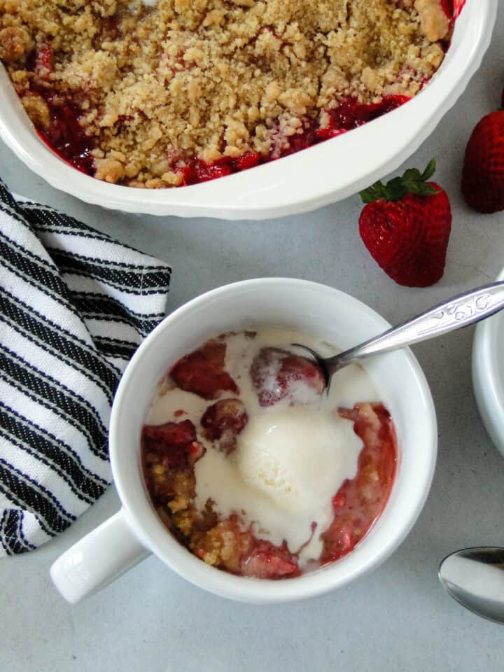 Strawberry crumble in white mug with spoon in front of dish of crumble.