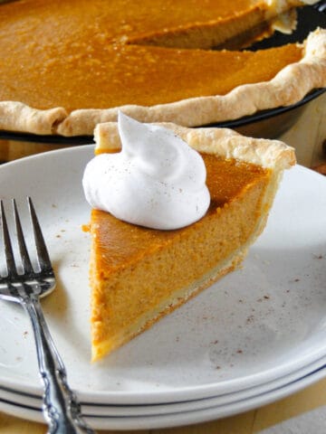 Front view of a slice of pumpkin pie topped with whipped cream on a white round plate and fork on the side.