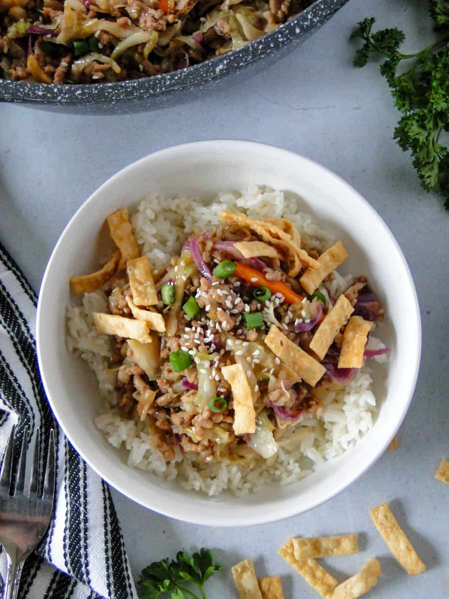 Top view of egg roll in a bowl served over white rice and garnished with fried wonton strips, green onion slices and sesame seeds with fork on the side.