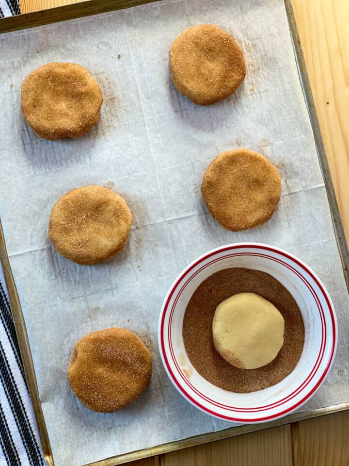 Cookie dough on parchment lined cookie sheet, with one cookie dough puck in bowl of cinnamon sugar mix.