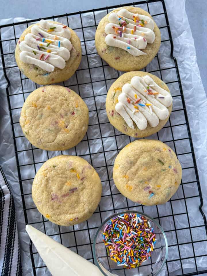 Birthday cake cookies on wire rack with 3 cookies frosted, a bowl of sprinkles and a bag of frosting.