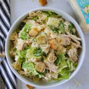 Chicken caesar pasta salad in white serving bowl with bottle of dressing to right side.