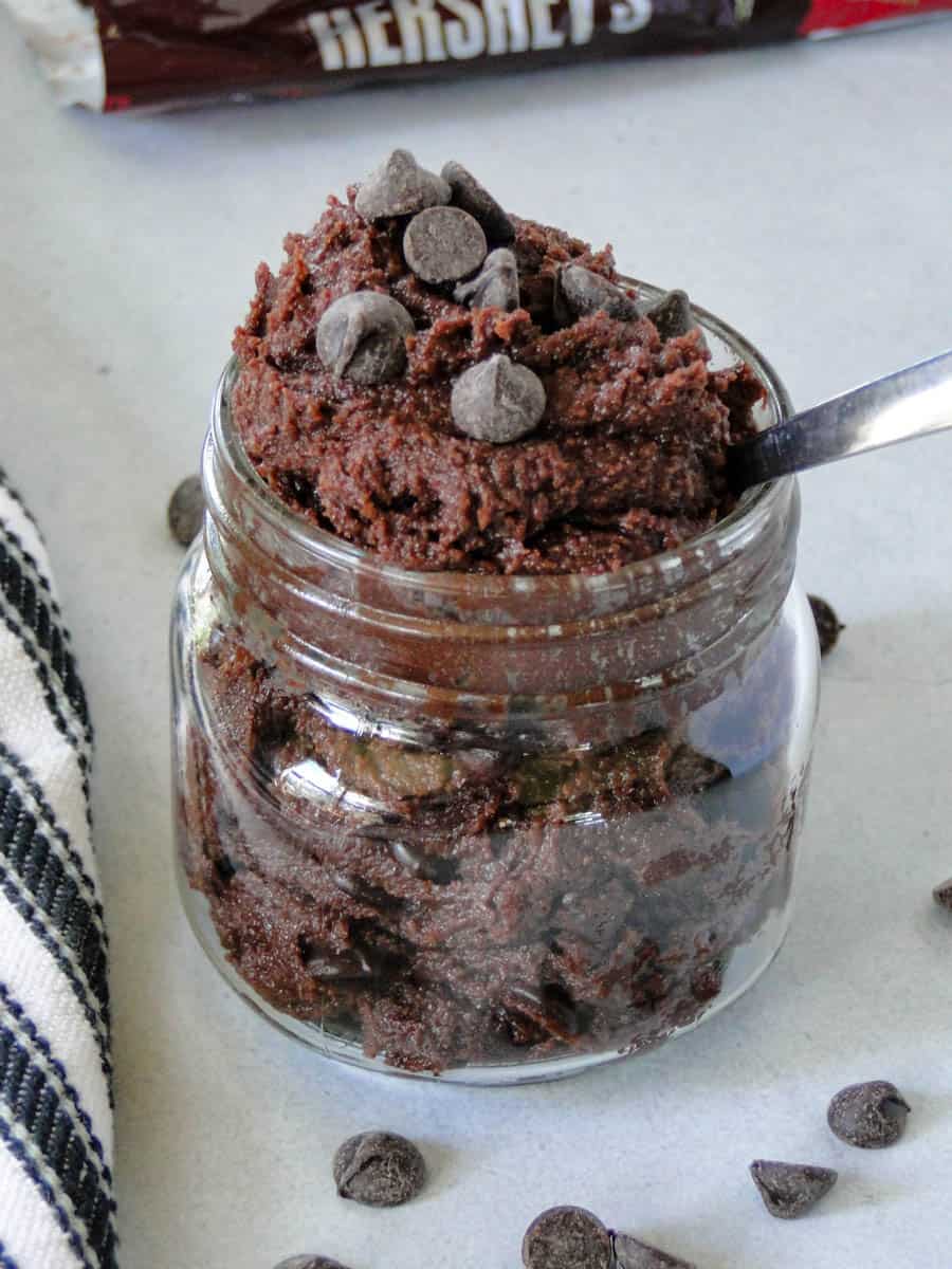 Edible brownie batter in mason jar topped with extra chocolate chips and with a spoon in jar.