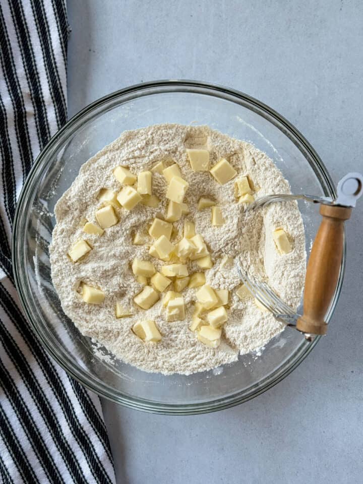 Pea sized butter cubes added to flour mixture in glass mixing bowl with pastry blender.