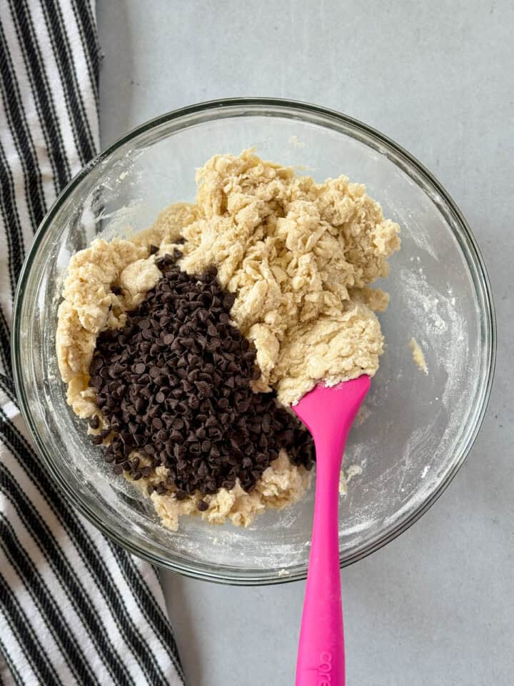 Mini chocolate chips added to scone dough in glass mixing bowl with pink spatula.