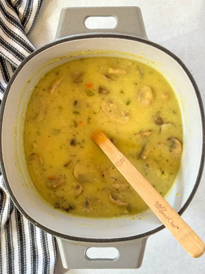 Chicken broth added to veggies in dutch oven with wooden spoon.