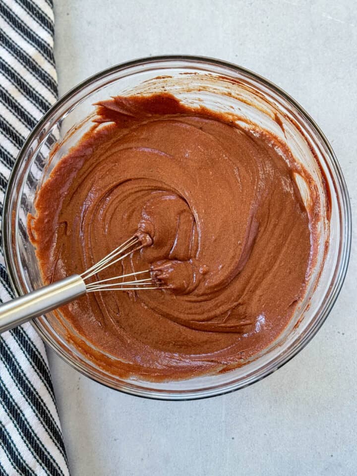 Eggs and vanilla extract whisked into chocolate mixture in glass mixing bowl with flat whisk.