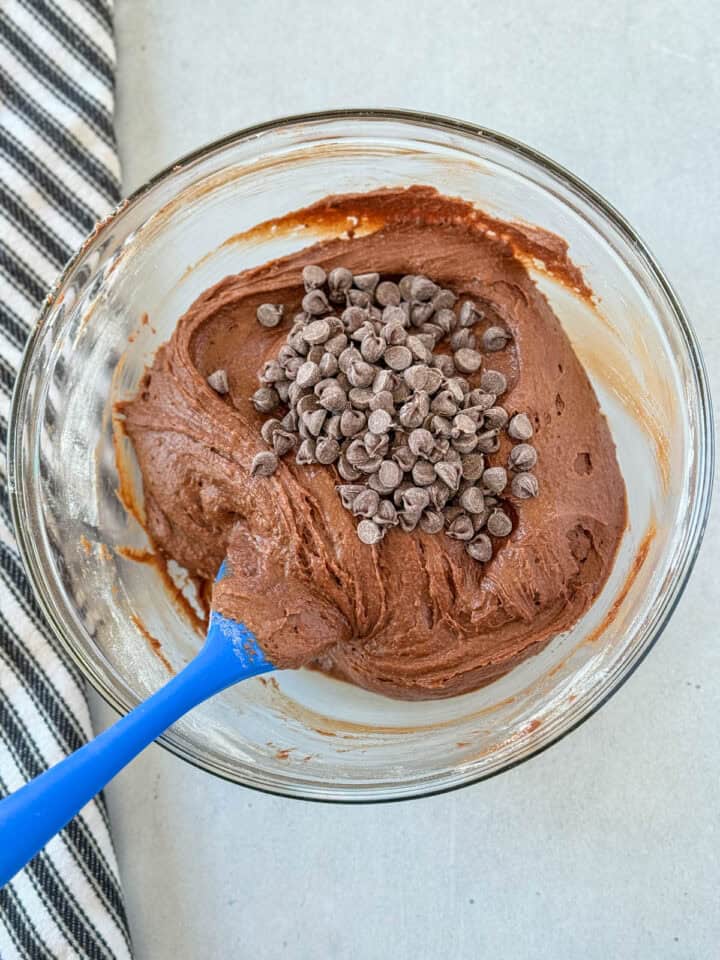 Chocolate chips added to brownie batter in glass mixing bowl with blue spatula.