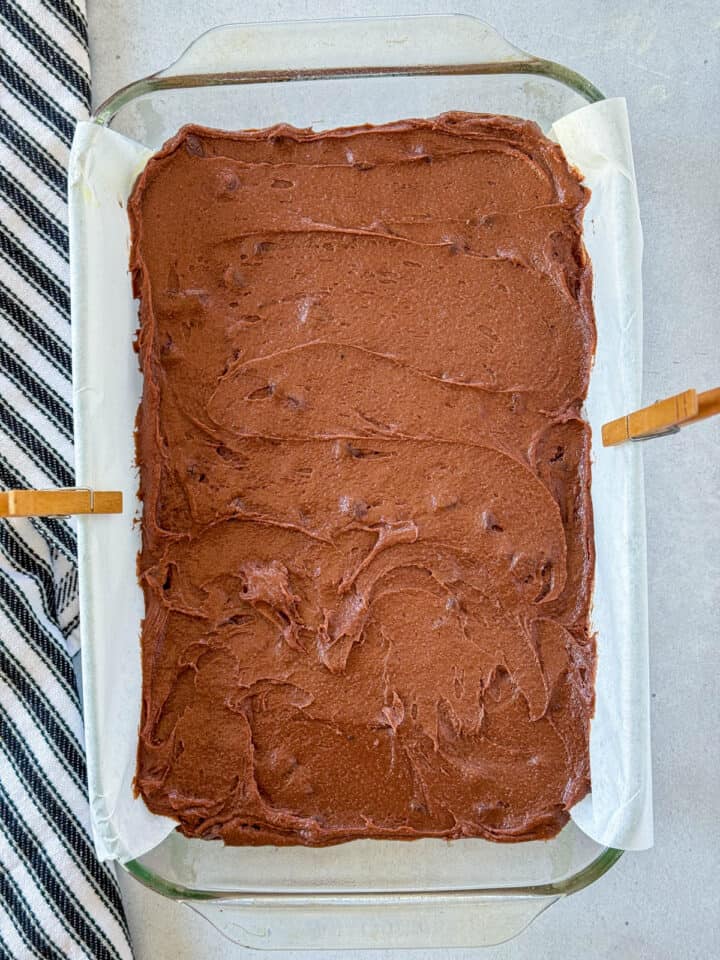 Brownie batter spread out in 9x13 glass baking dish lined with parchment paper.